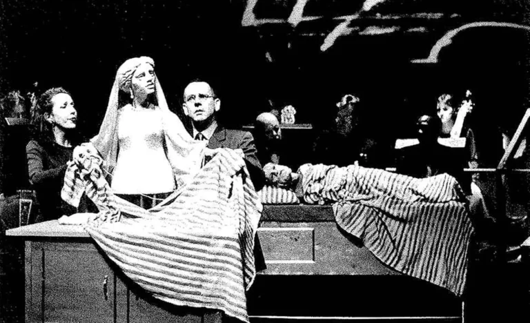 A l’avant-plan Pénélope file la laine dans la couverture-costume d’Ulysse Guillaumette Laurens, chanteuse, Adrian Kohler, acteur-manipulateur. À l’arrière-plan Ulysse, alité est soutenu par Scott Weir, chanteur, Basil Jones, acteur-manipulateur et les musiciens. Photo Herman Sorgeloos.